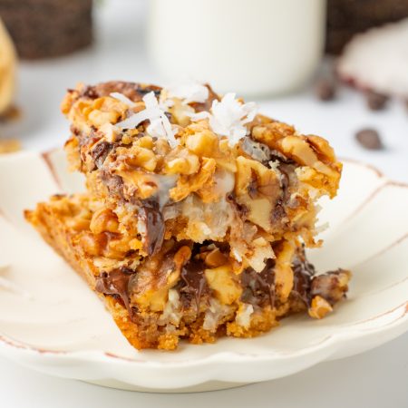 Two dessert bars with chocolate chips, nuts, and shredded coconut are stacked on a white plate.