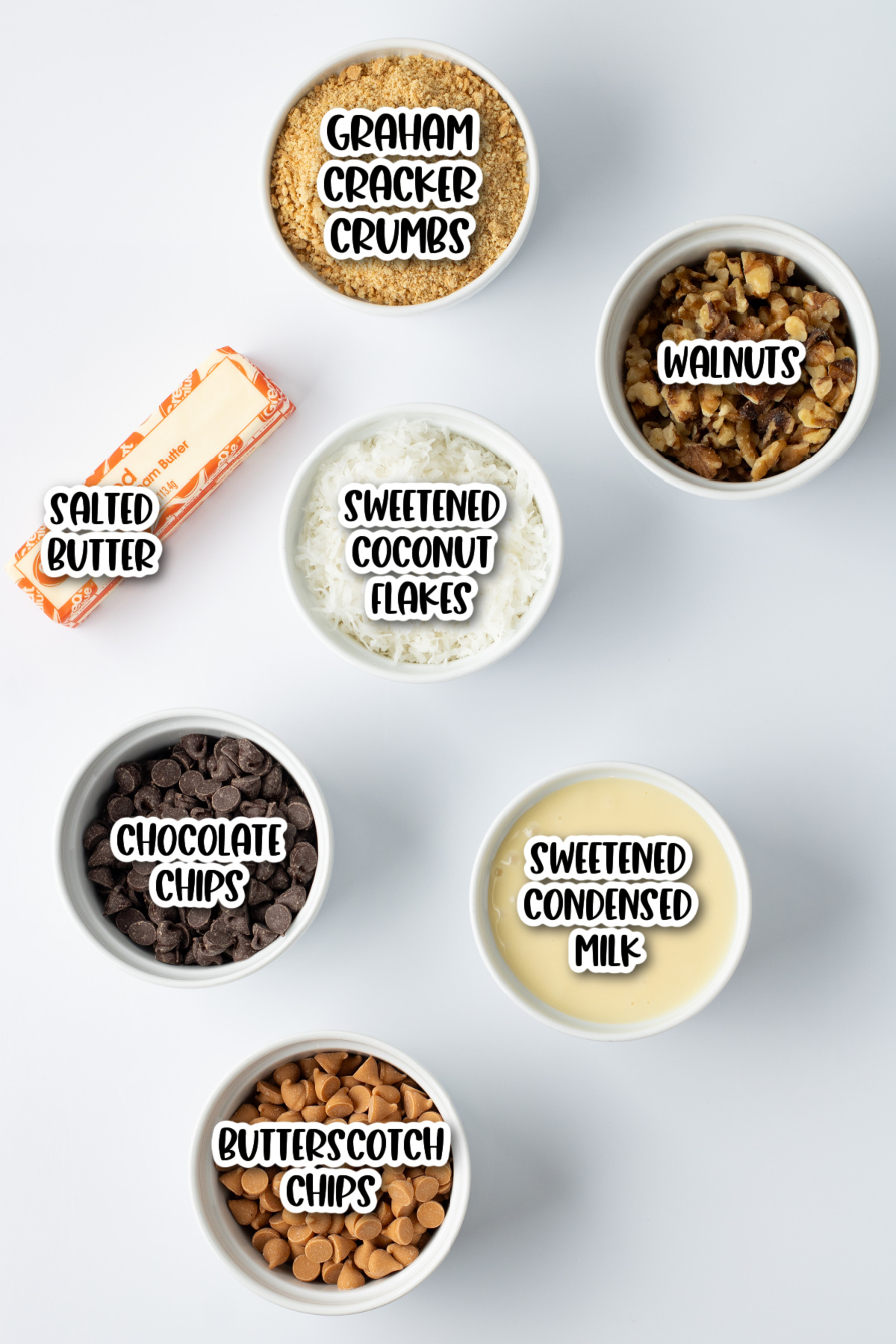 Seven baking ingredients displayed in bowls, including graham cracker crumbs, walnuts, sweetened coconut flakes, salted butter, chocolate chips, butterscotch chips, and sweetened condensed milk.