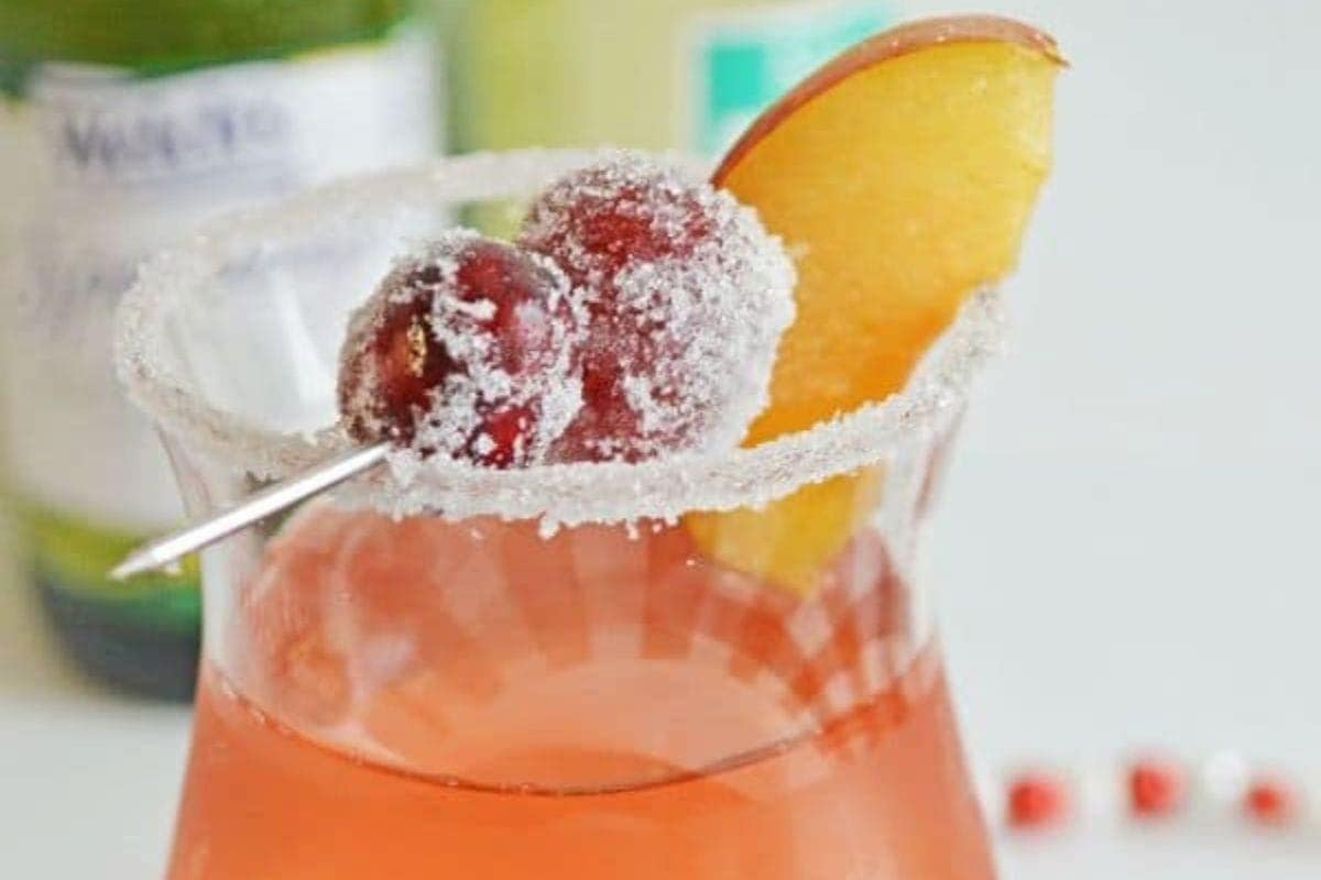 A glass of pink cocktail garnished with a sugared rim, two sugared cranberries on a pick, and a peach slice.