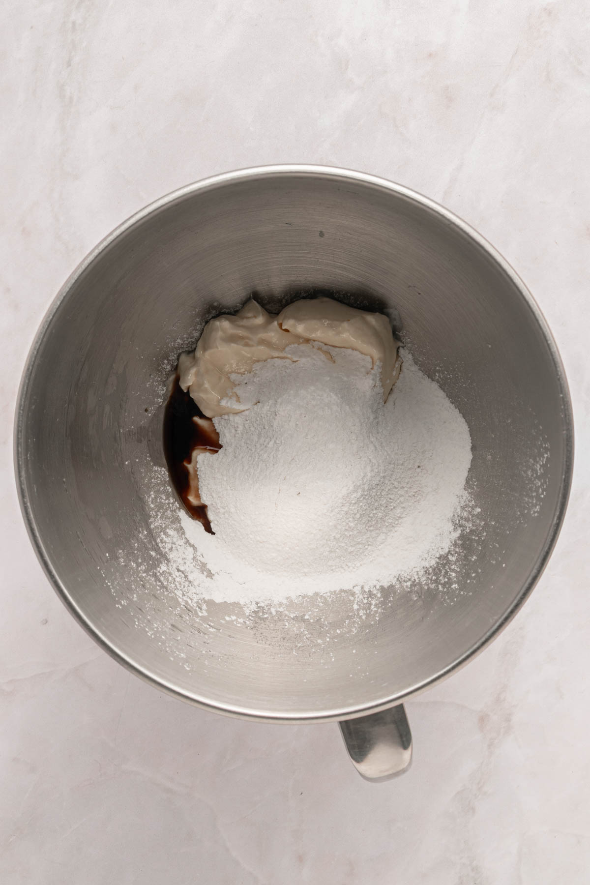 A metal mixing bowl containing powdered sugar, cheese, and vanilla extract on a light-colored surface.