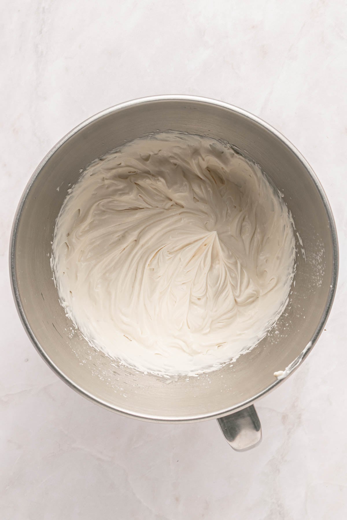 A stand mixer bowl filled with freshly whipped, smooth white frosting, viewed from above on a light-colored surface.