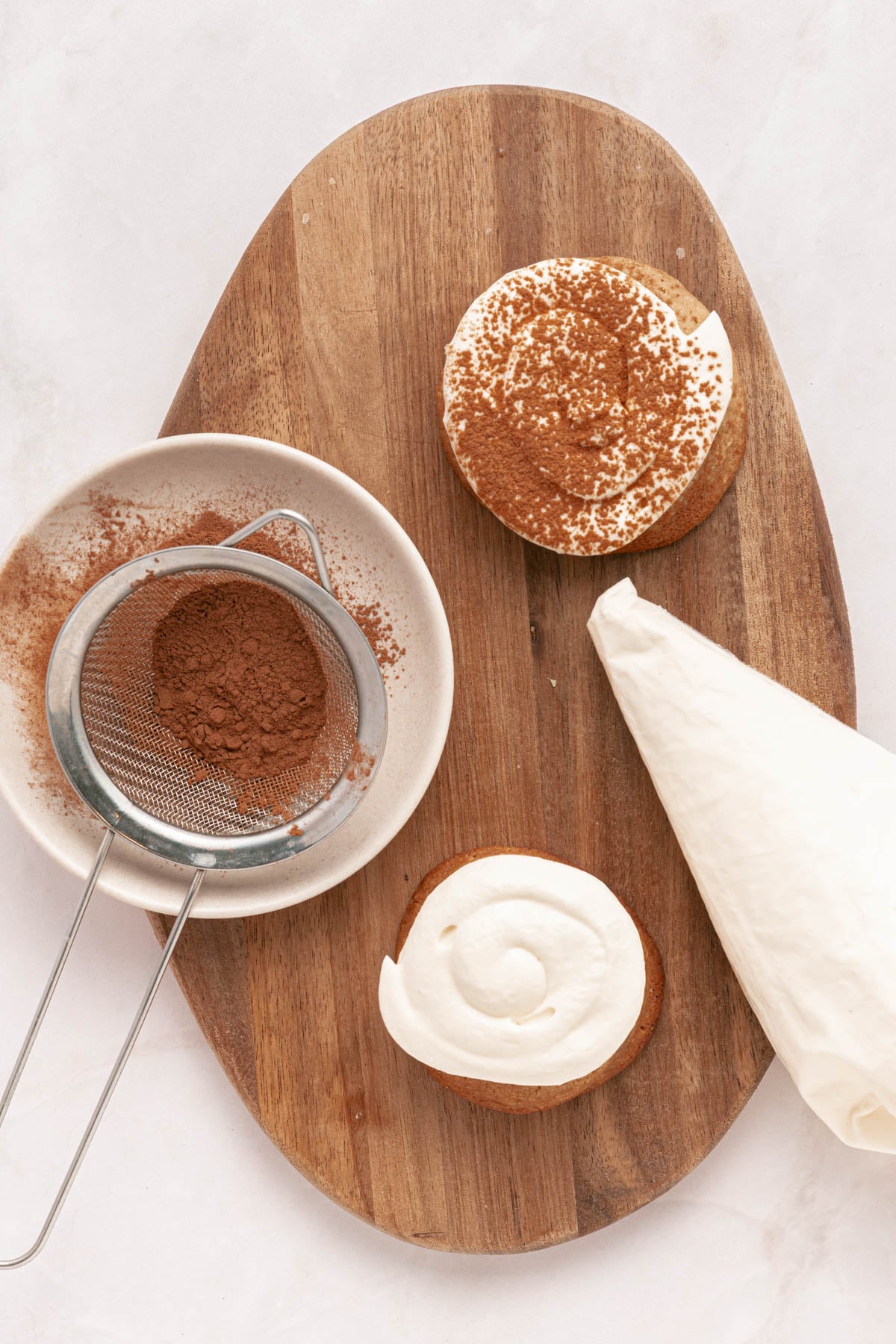 A plate with a fine-mesh strainer holding cocoa powder, two cookies with piped cream, and a pastry bag of cream on a wooden board.