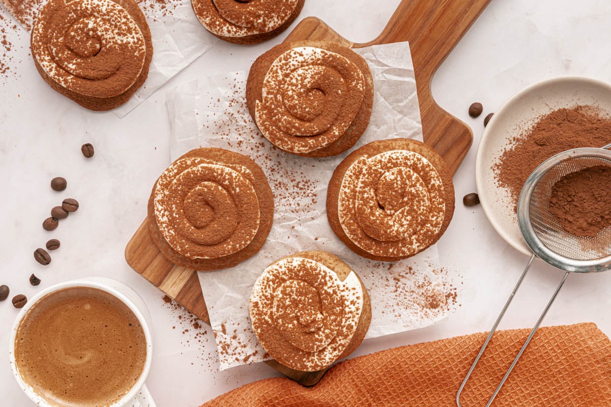 Six swirled tiramisu cookies dusted with cocoa powder on a wooden board, surrounded by a cup of coffee, a sieve, and scattered coffee beans.