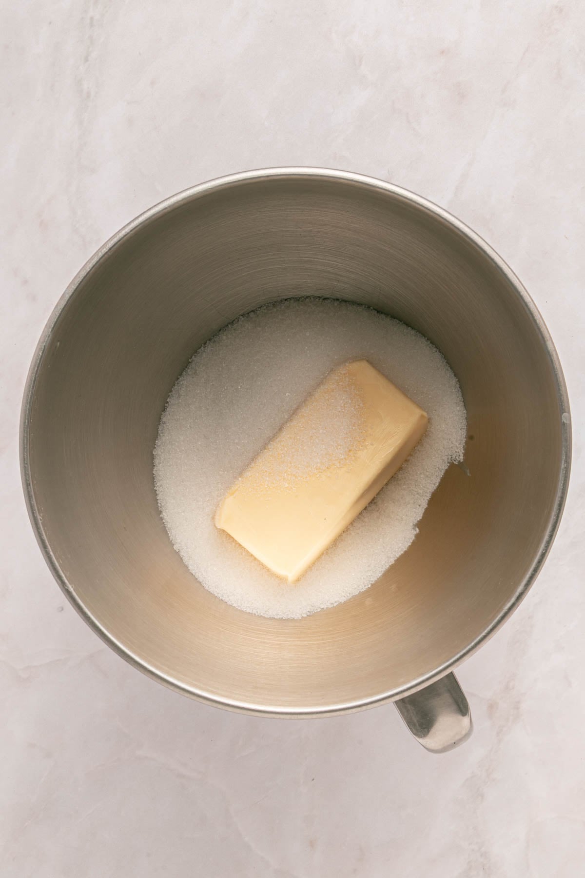 A metal mixing bowl containing a stick of butter and granulated sugar on a light-colored surface.