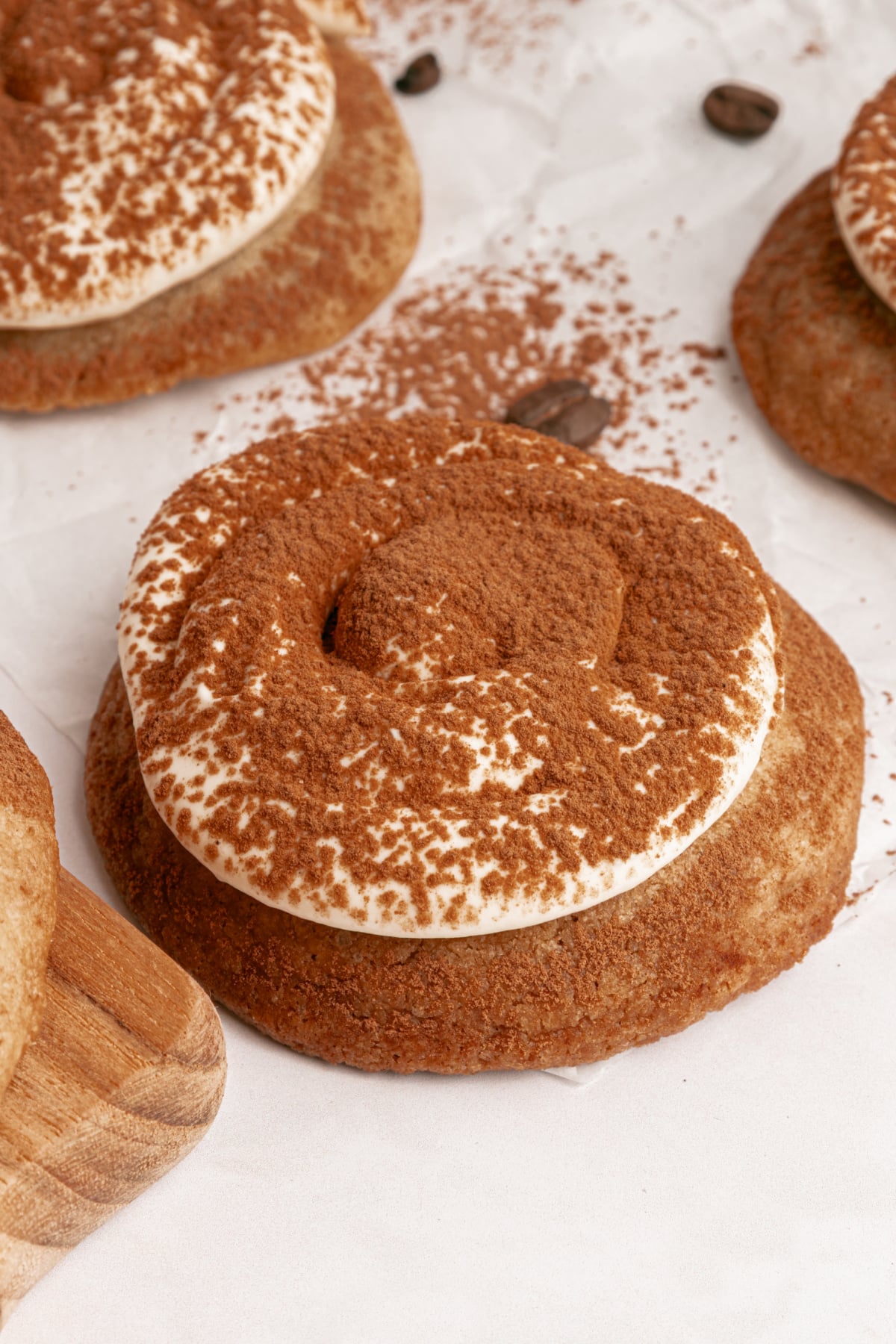 A close-up of a cookie topped with cream and dusted with cocoa powder, placed on parchment paper with other cookies and coffee beans nearby.