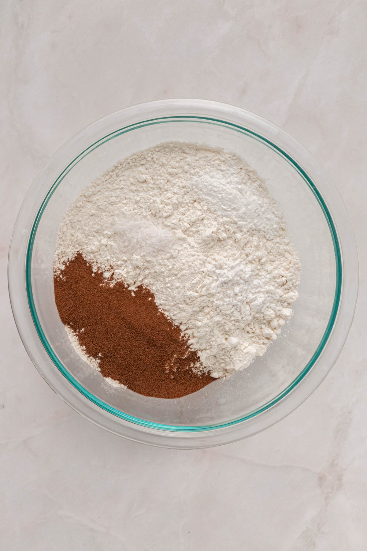 A glass bowl containing flour, cinnamon, and baking powder, displayed on a light-colored surface.