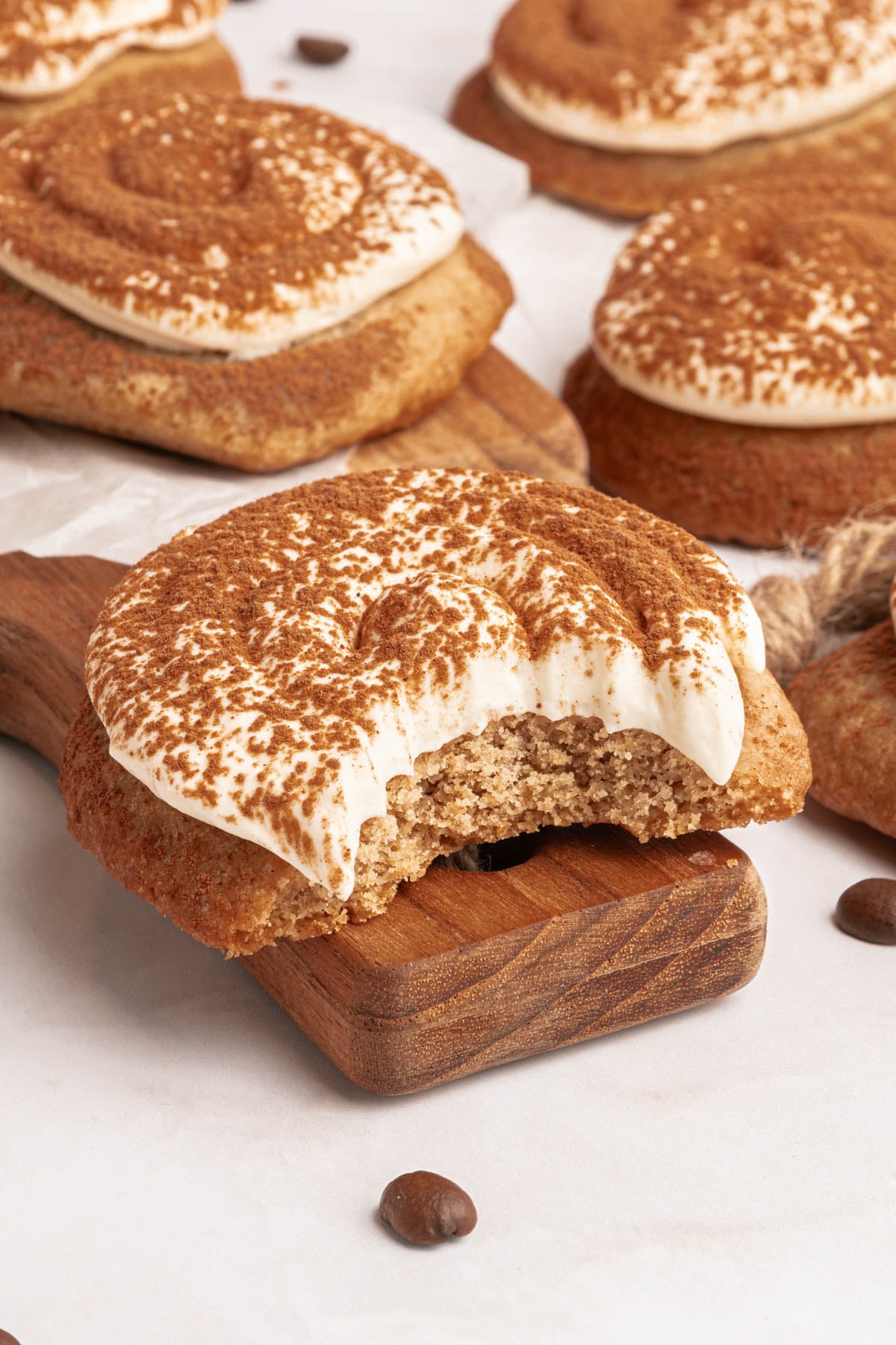 A coffee-flavored cookie with white frosting and cocoa powder, showing a bite taken out, on a wooden board with whole cookies in the background.