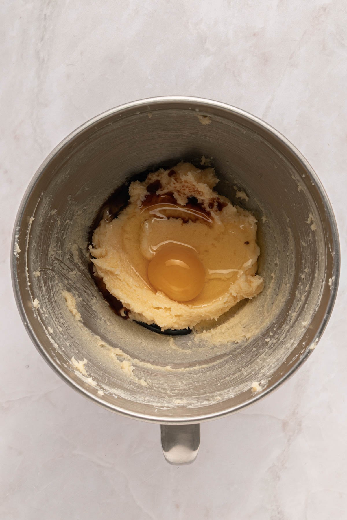 A metal mixing bowl containing creamed butter and sugar with a cracked egg and vanilla extract on top, ready to be mixed.