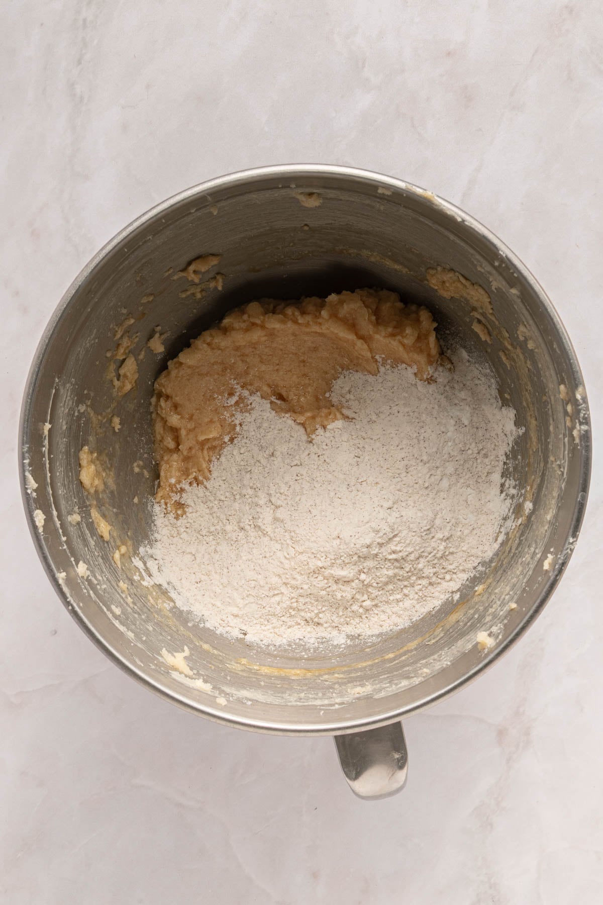 A mixing bowl contains partially mixed dough with flour on top, sitting on a light-colored countertop.