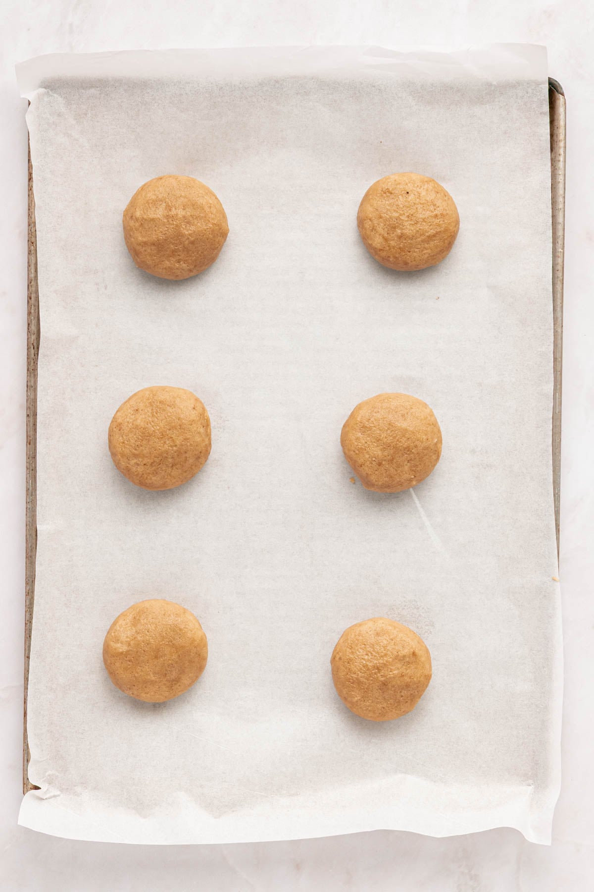 Six raw cookie dough balls are evenly spaced on a parchment-lined baking sheet, ready to be baked.
