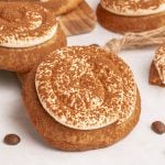 A large cookie topped with swirled cream frosting and dusted with cocoa powder, placed on a white surface with coffee beans nearby.