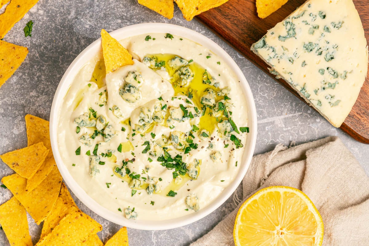 A bowl of creamy dip topped with crumbled blue cheese, herbs, and olive oil, surrounded by tortilla chips, a wedge of blue cheese, and a halved lemon.