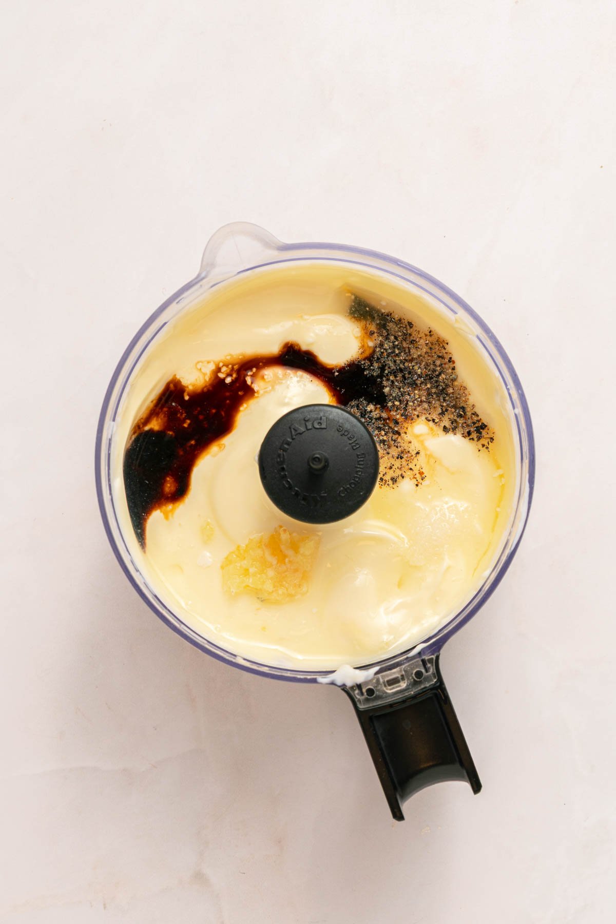 A food processor bowl containing mayonnaise, Worcestershire sauce, black pepper, and minced garlic on a white surface.