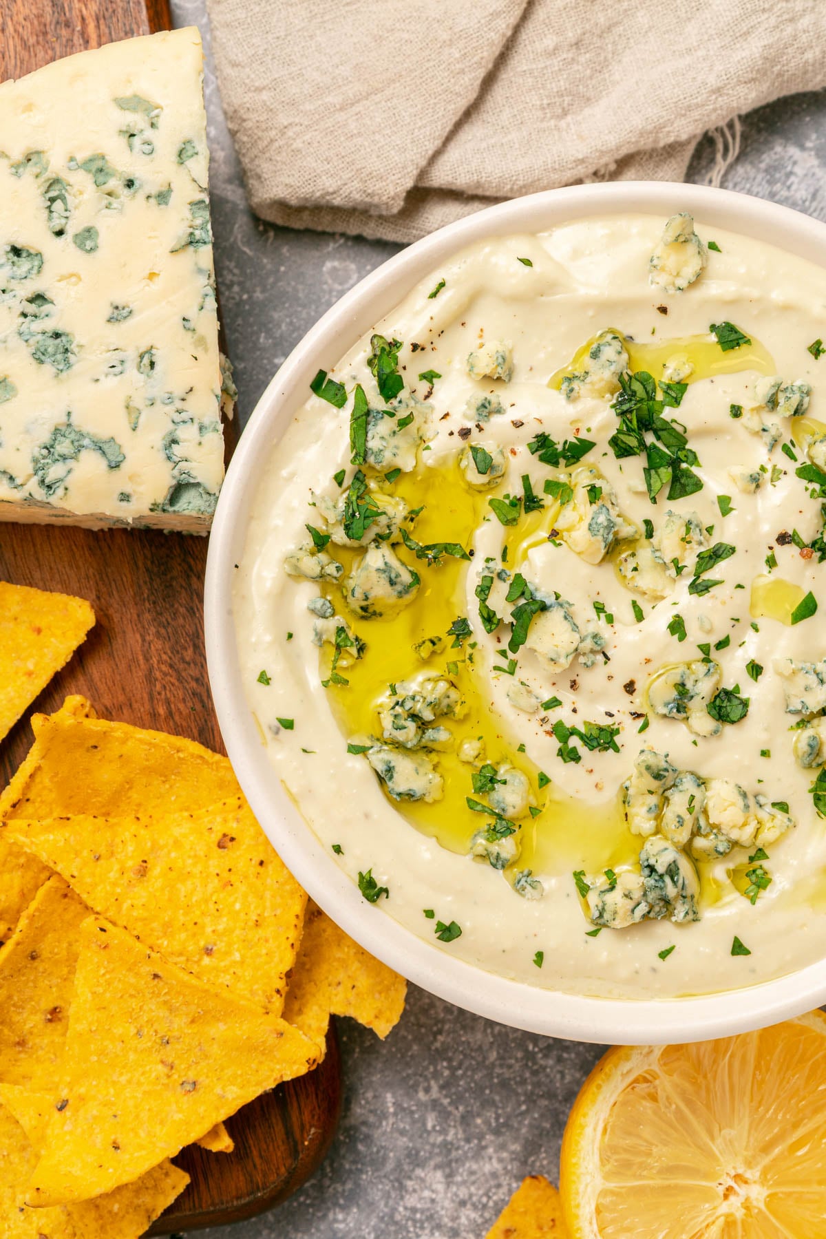 A bowl of creamy blue cheese dip garnished with blue cheese, herbs, and olive oil, surrounded by tortilla chips, a wedge of blue cheese, and a half lemon on a gray surface.
