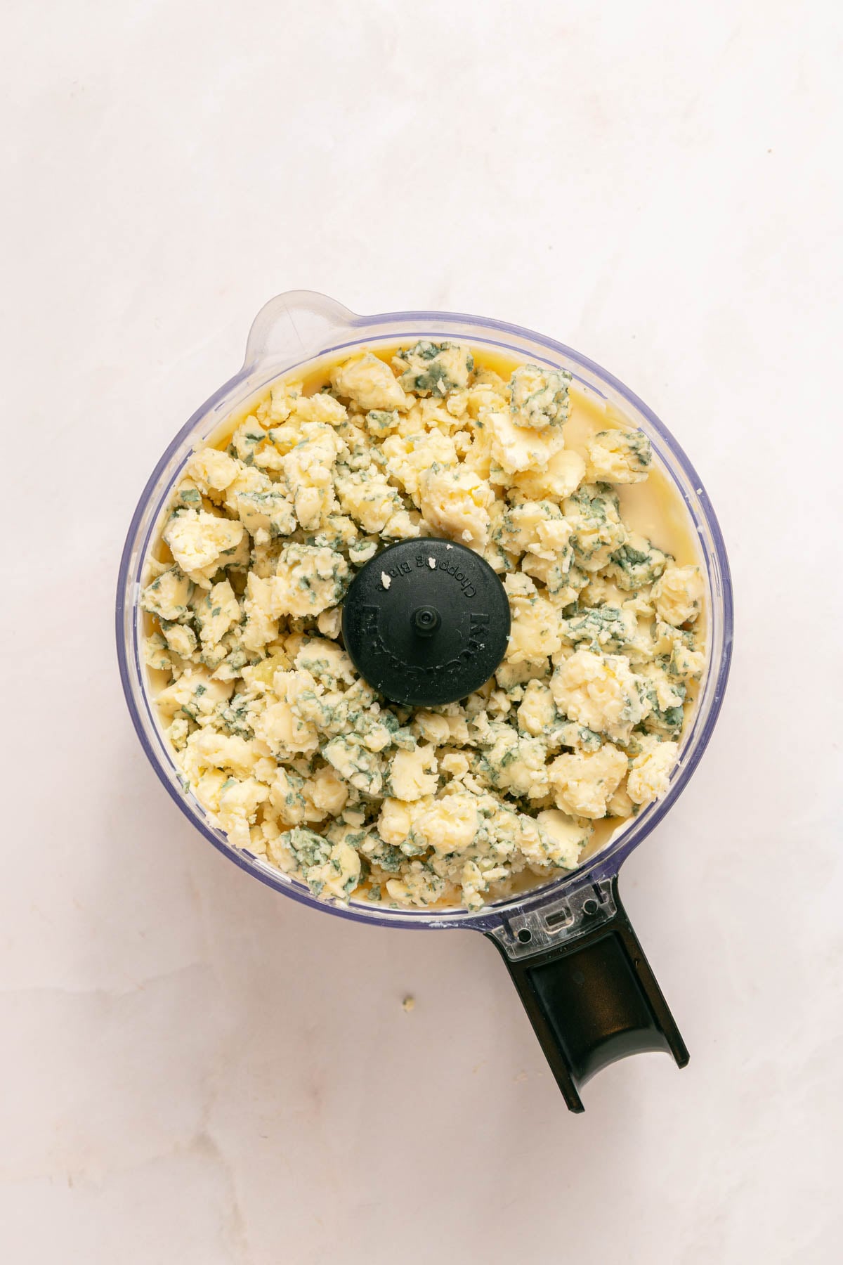 A food processor bowl filled with crumbled blue cheese and cream cheese mixture on a light-colored surface.