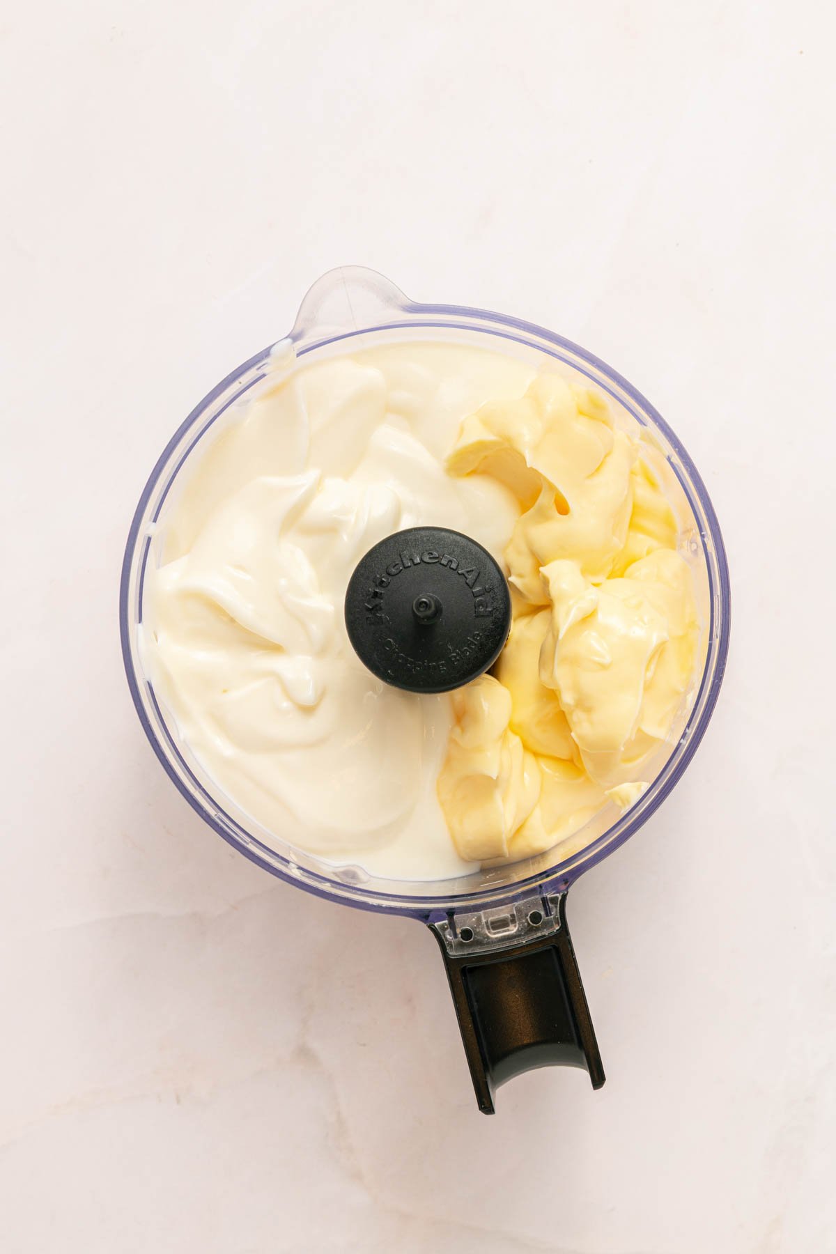 A food processor bowl containing white mayonnaise on one side and sour cream on the other, viewed from above.