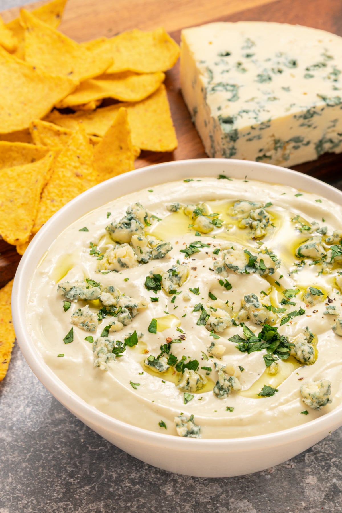 A bowl of creamy dip topped with crumbled blue cheese and herbs, with tortilla chips and a block of blue cheese on a wooden board in the background.