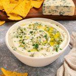 A bowl of creamy dip garnished with herbs and blue cheese crumbles, surrounded by tortilla chips and a block of blue cheese in the background.