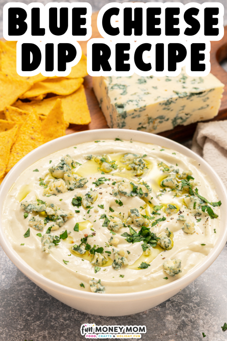 A bowl of blue cheese dip garnished with herbs and crumbled blue cheese, surrounded by tortilla chips and a block of blue cheese.