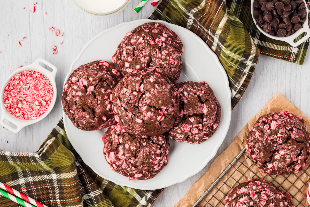 A plate of chocolate cookies with crushed peppermint pieces, surrounded by a glass of milk, plaid napkins, chocolate chips, and additional peppermint on a light wooden surface.