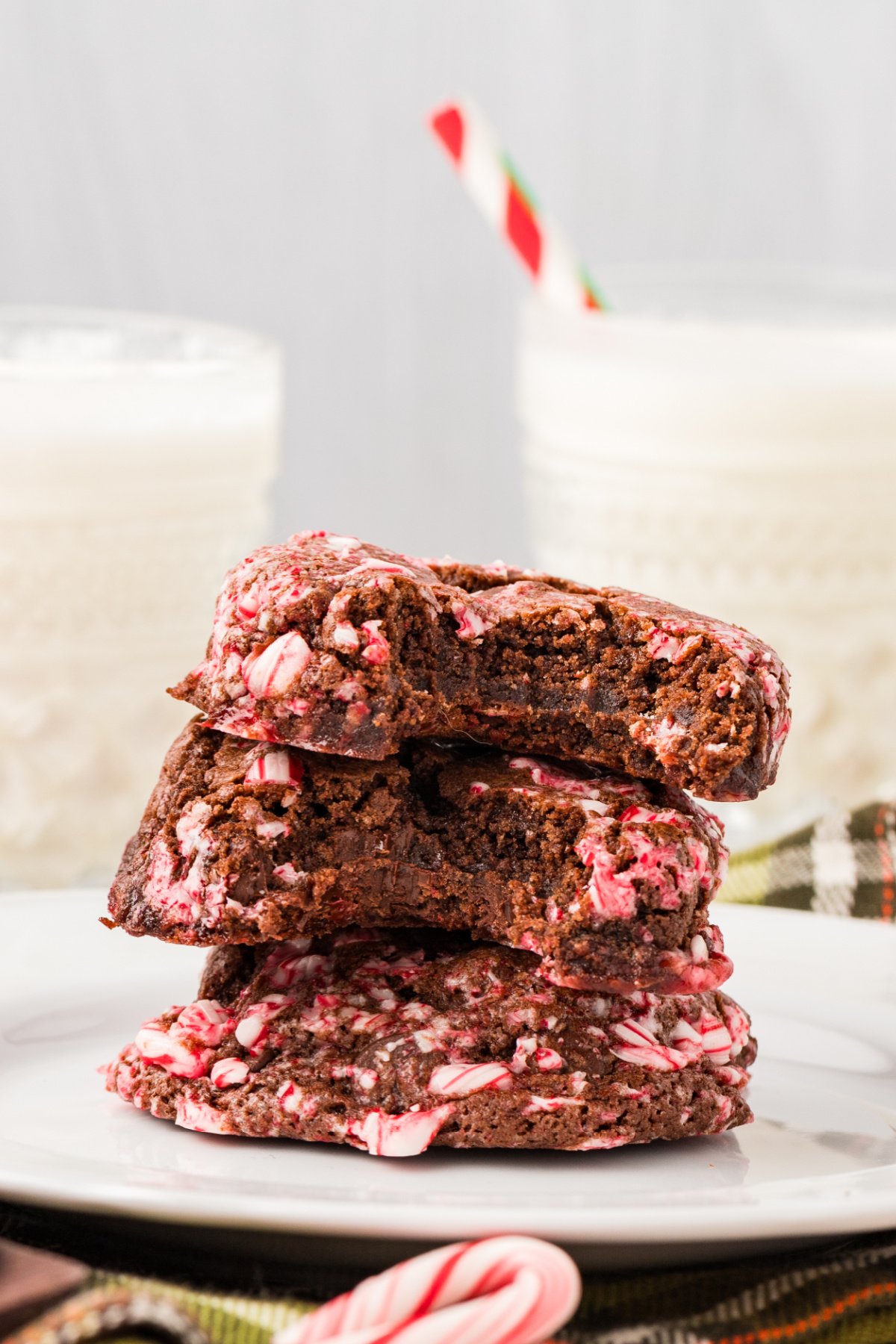 A stack of three chocolate cookies topped with crushed peppermint candy sits on a white plate, with glasses of milk and a candy cane in the background.