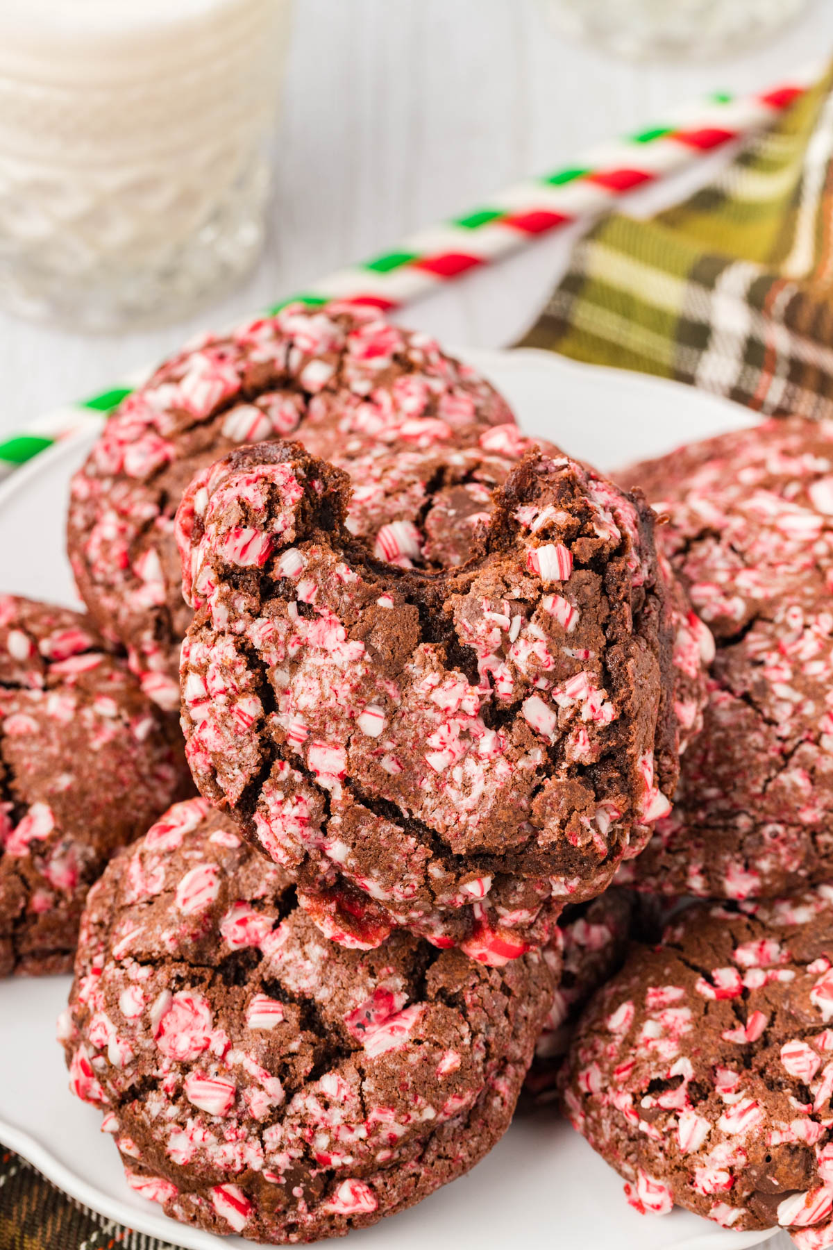 A plate of chocolate cookies topped with crushed peppermint candy, with one cookie showing a bite taken out. A glass of milk and striped straw are in the background.