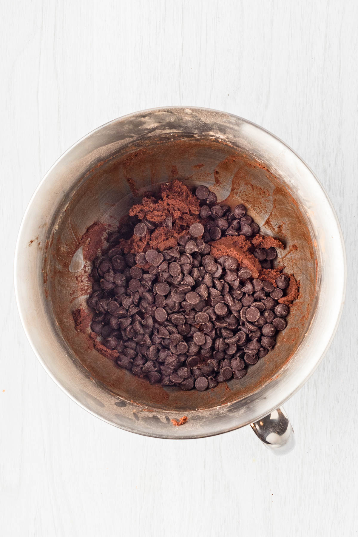 A metal mixing bowl containing cocoa powder and chocolate chips on a white surface.