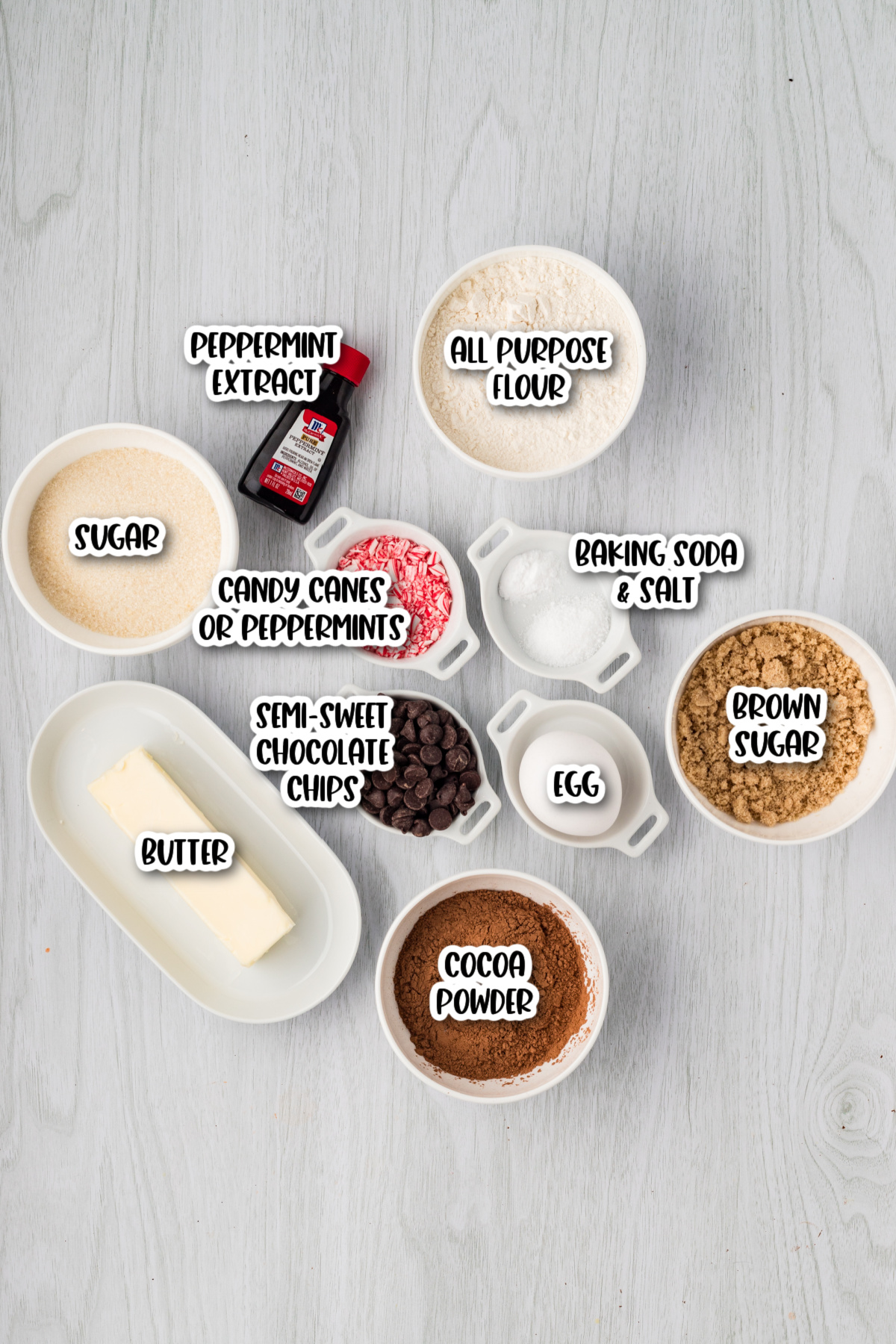 Assorted baking ingredients including butter, sugar, brown sugar, egg, chocolate chips, cocoa powder, flour, crushed candy canes, peppermint extract, baking soda, and salt.