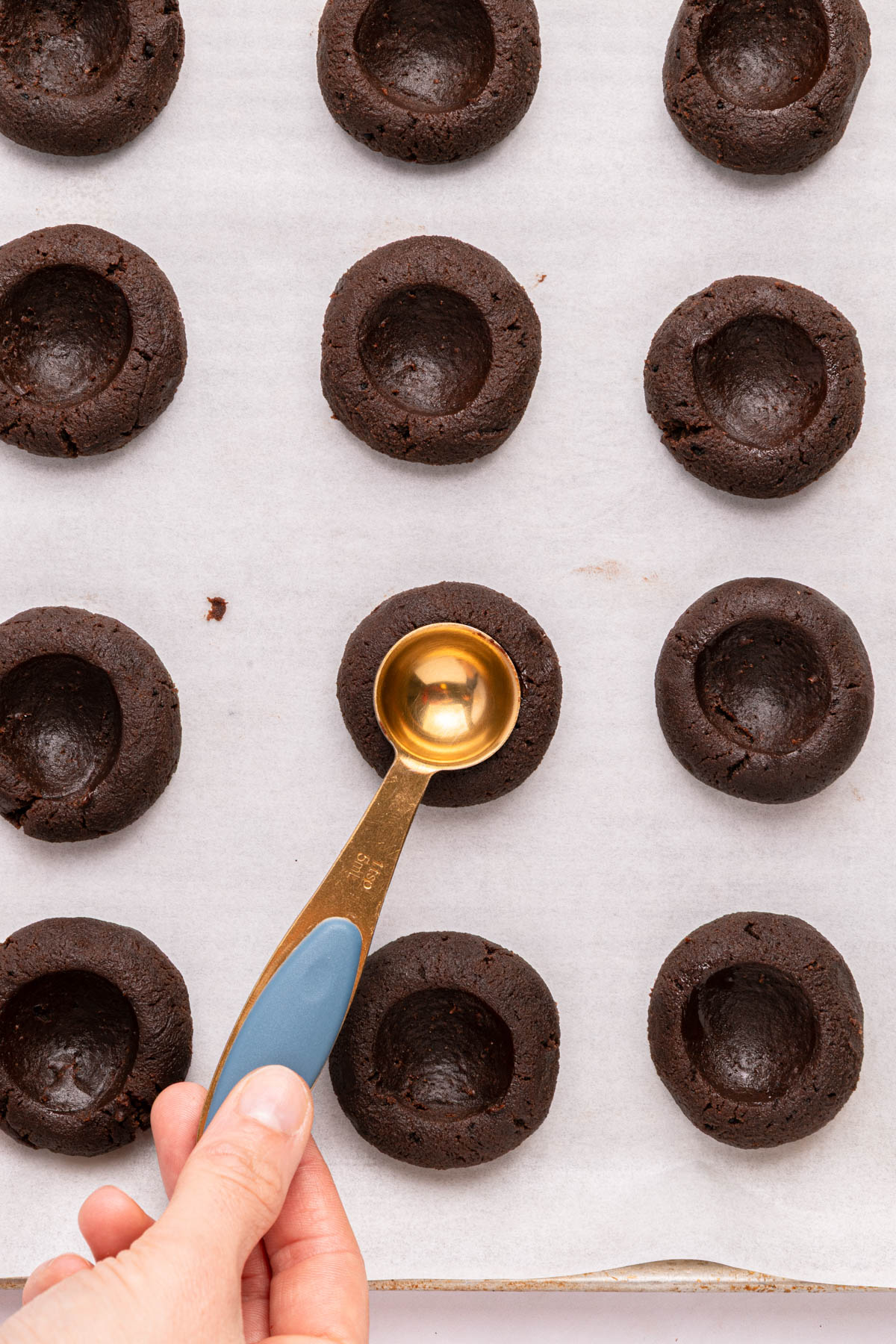 A hand uses a round measuring spoon to press indentations into chocolate cookie dough balls arranged on a parchment-lined baking sheet.