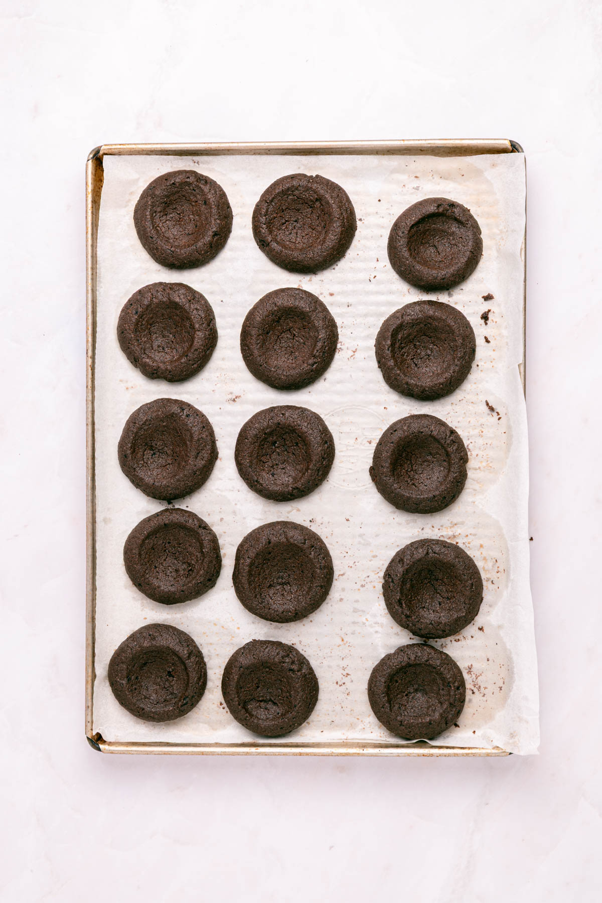 A baking tray lined with parchment paper holds 15 round chocolate cookies, each with an indented center.