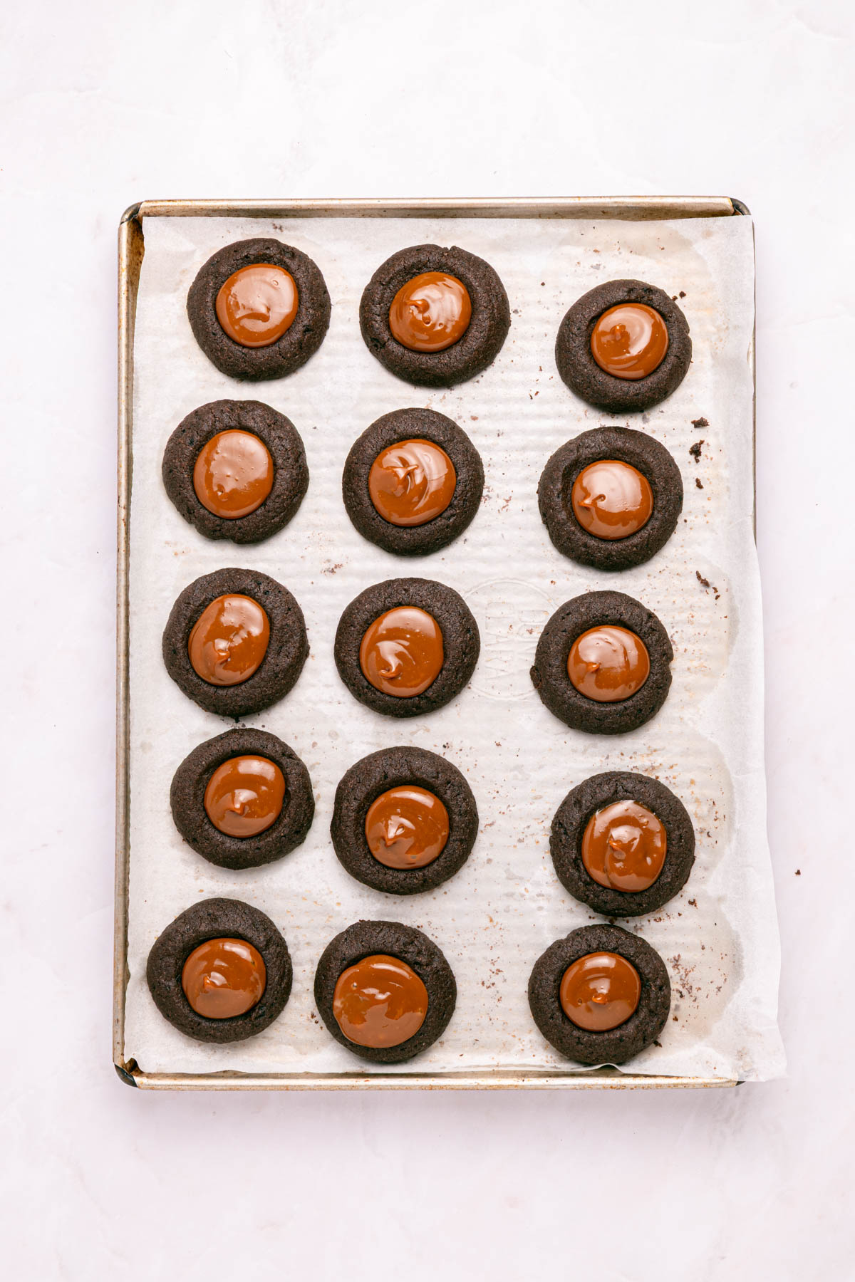 A baking tray with fifteen chocolate thumbprint cookies filled with melted chocolate, arranged in rows on parchment paper.
