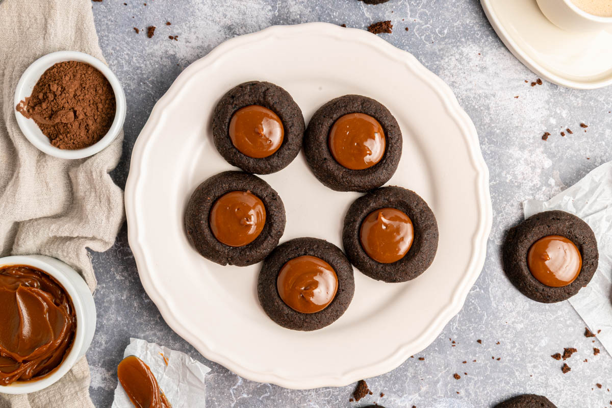 A white plate with six chocolate thumbprint cookies filled with caramel, surrounded by bowls of cocoa powder and caramel on a light gray surface.