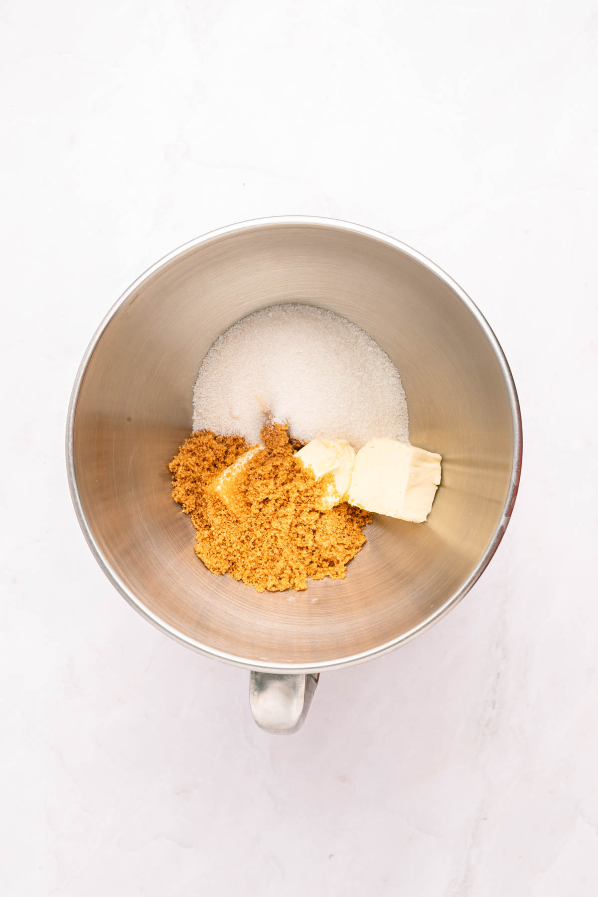 A metal mixing bowl containing white sugar, brown sugar, and two pieces of butter on a white surface.