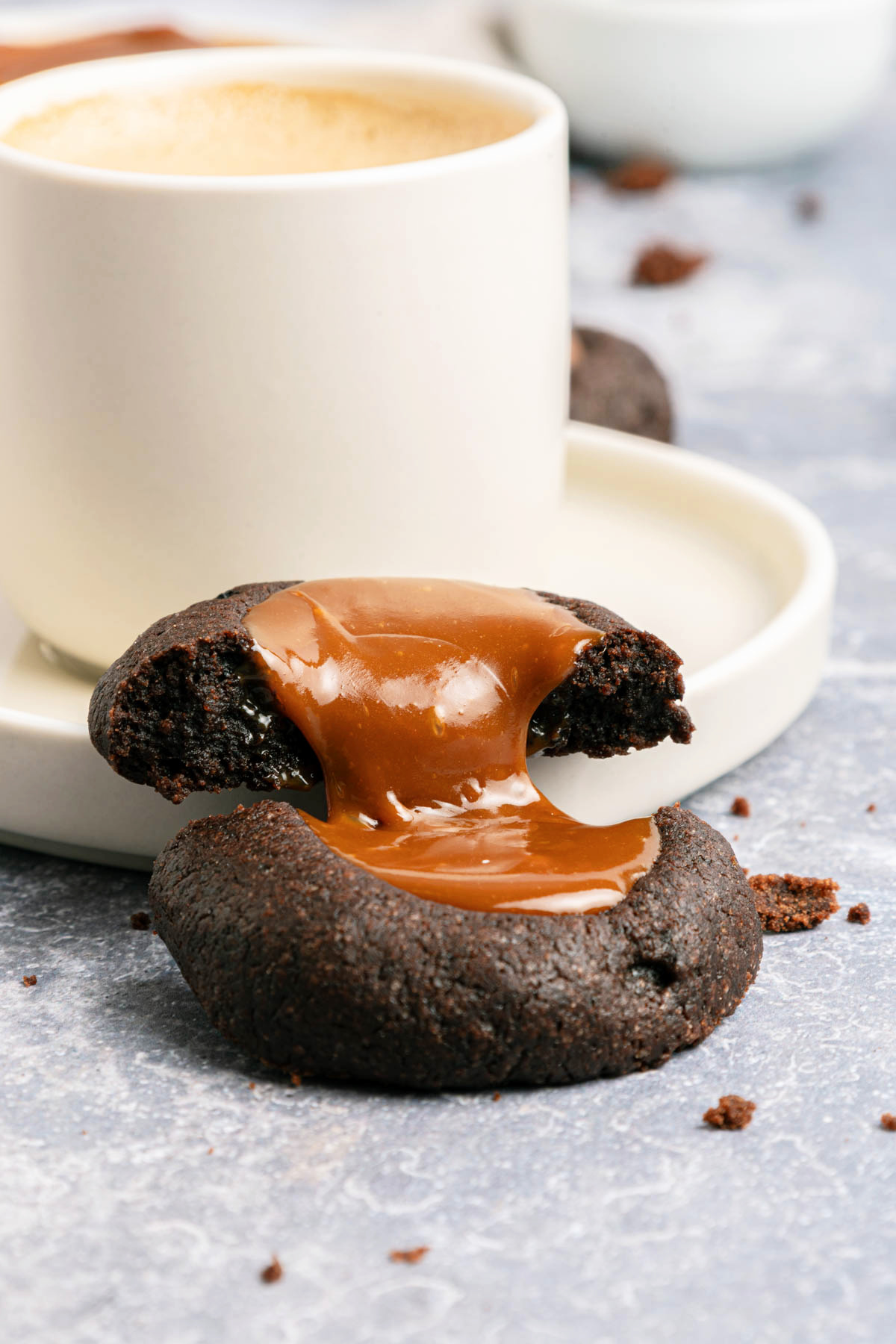 A chocolate cookie with a caramel center sits in front of a white cup of coffee on a saucer.