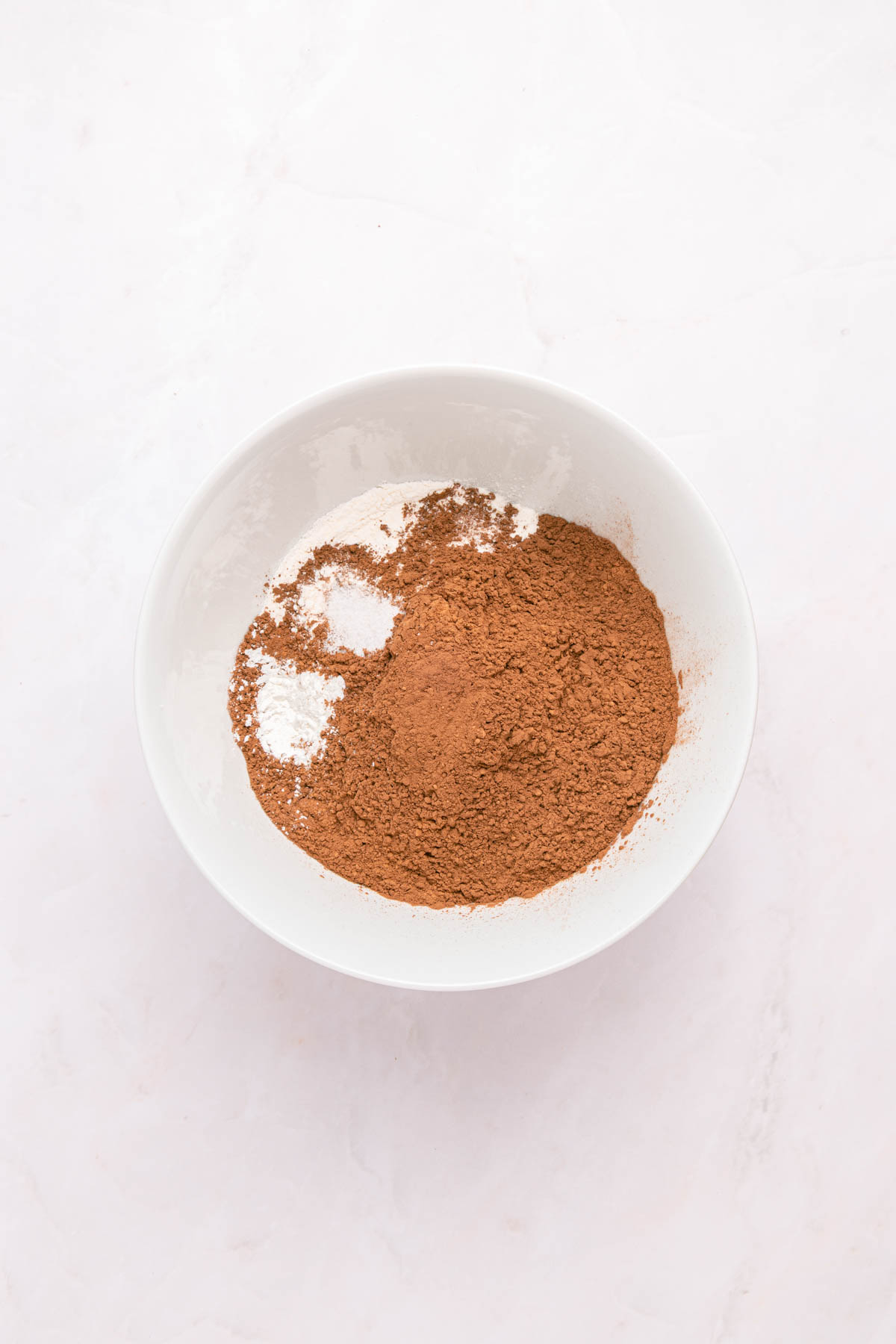 A white bowl containing a mixture of brown cocoa powder and a small amount of white baking soda on a light surface.