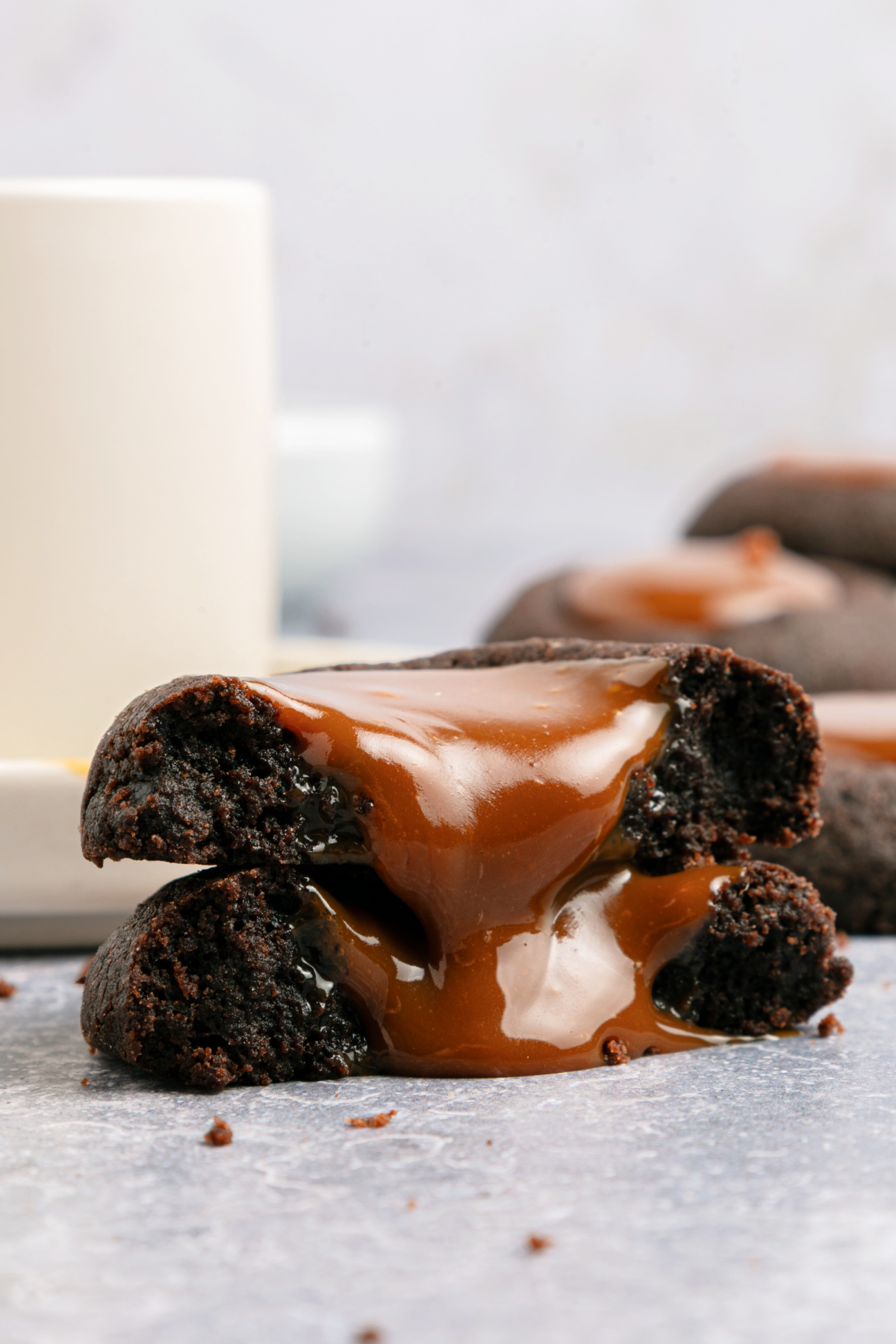 A close-up of a halved chocolate cookie with thick caramel filling oozing out, set on a smooth surface with a blurred cup in the background.