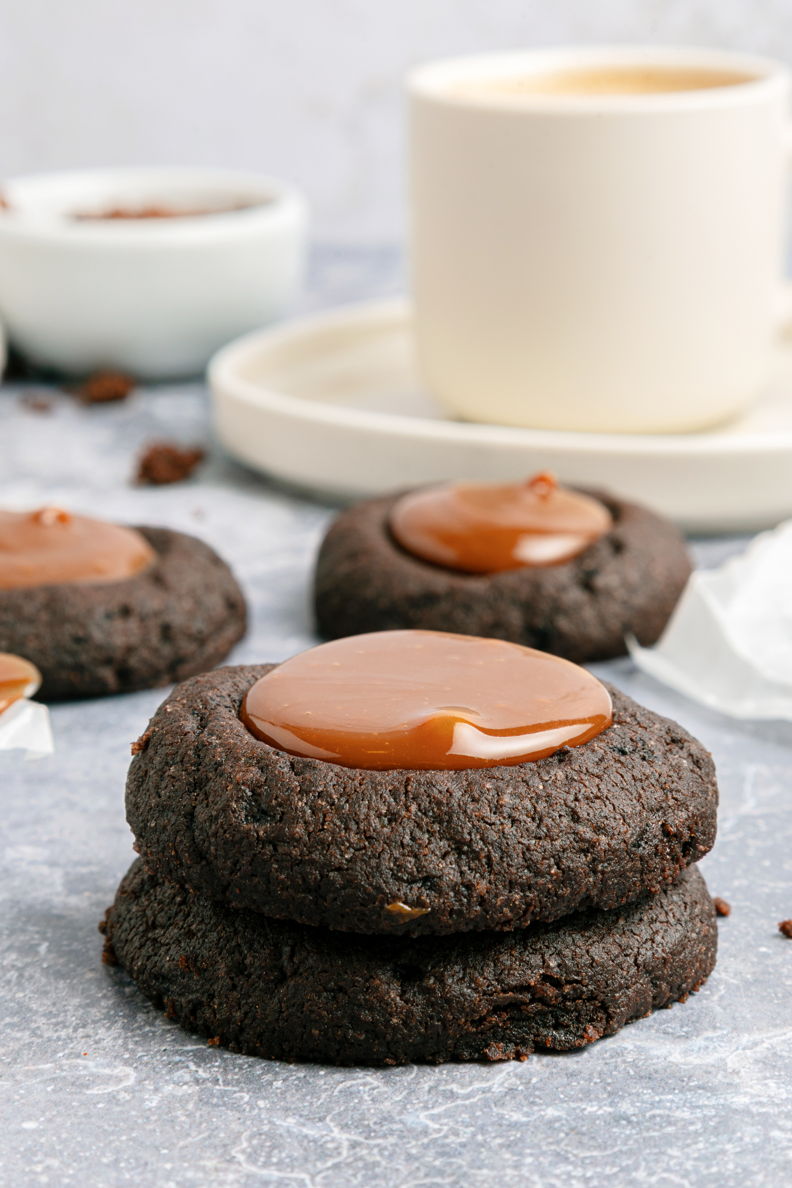 Two chocolate thumbprint cookies with caramel centers are stacked on a gray surface, with a cup of coffee and more cookies in the background.