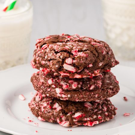 Three chocolate cookies with crushed peppermint pieces are stacked on a white plate, with glasses of milk in the background.
