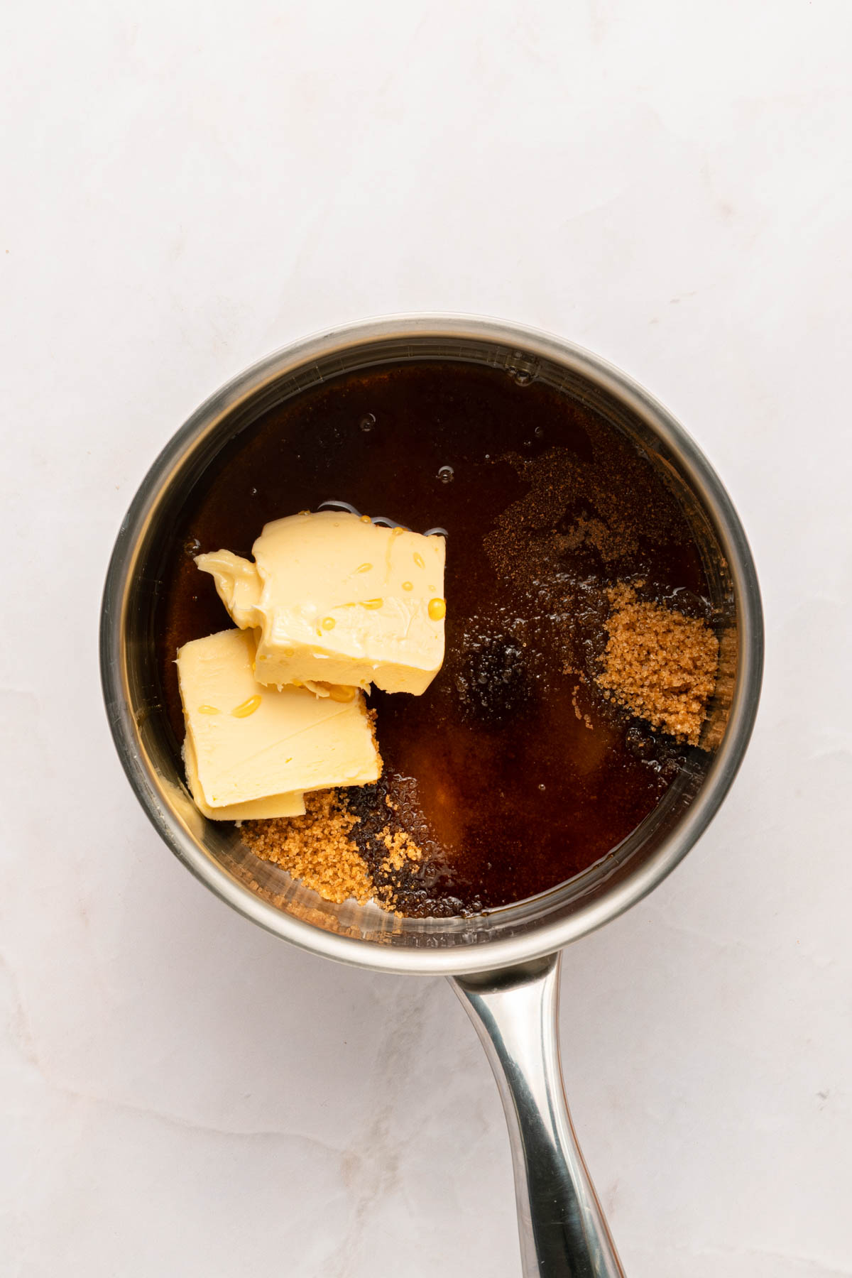 A saucepan containing brown sugar, butter, and a dark liquid, possibly molasses or syrup, on a light-colored surface.