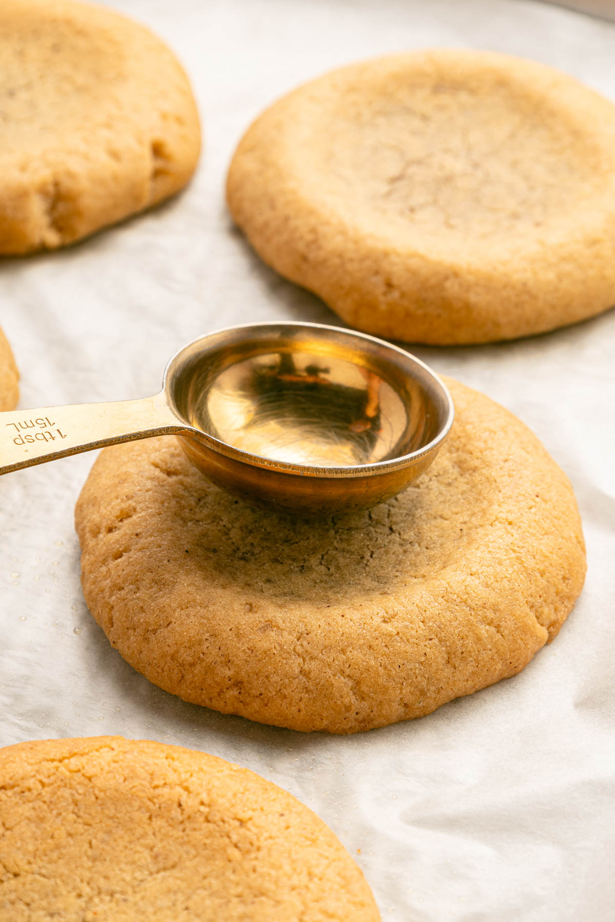 A metal tablespoon measuring spoon rests on a round cookie with an indentation, surrounded by other similar cookies on parchment paper.