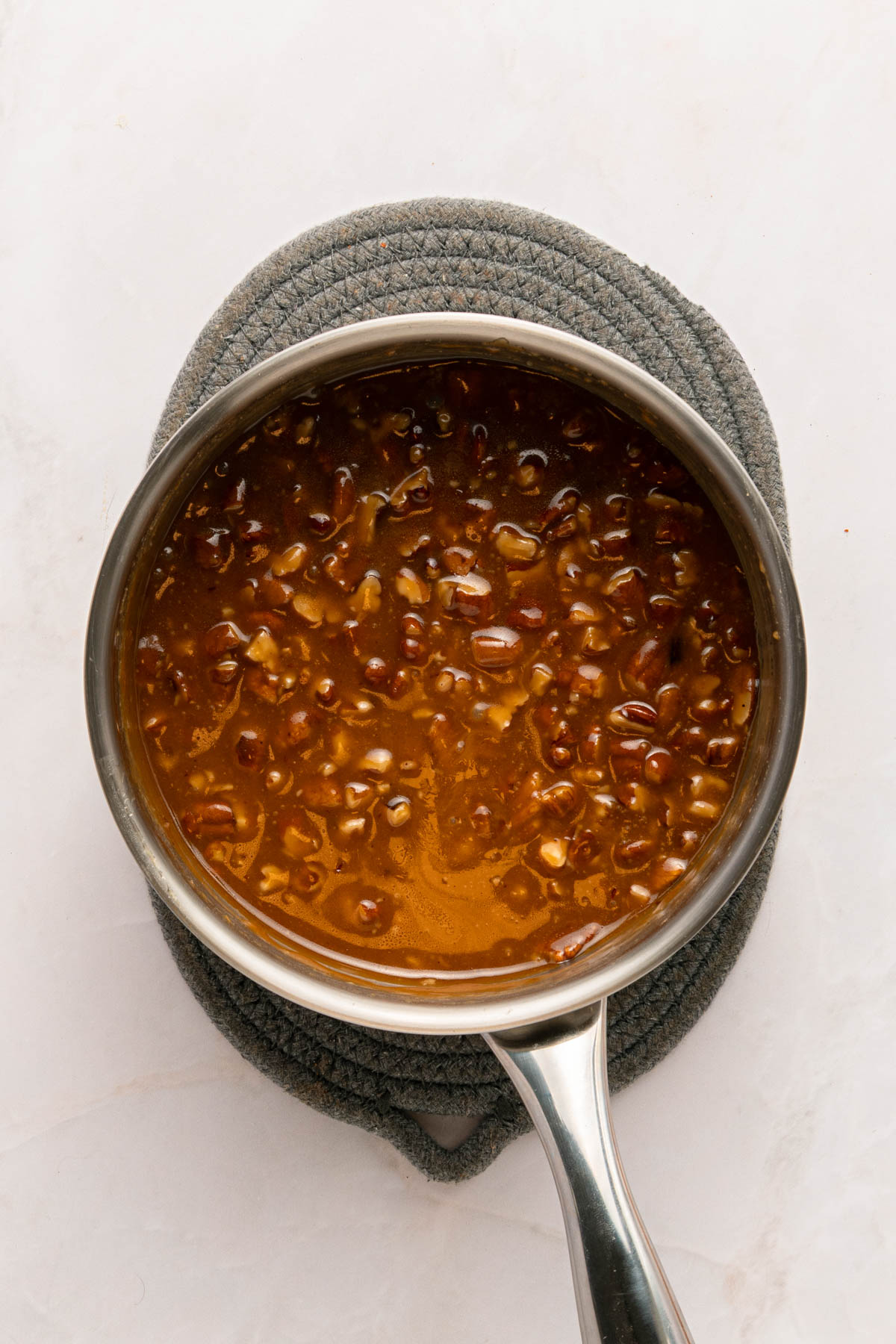 A stainless steel saucepan filled with  sauce sits on a textured gray pot holder against a light background.
