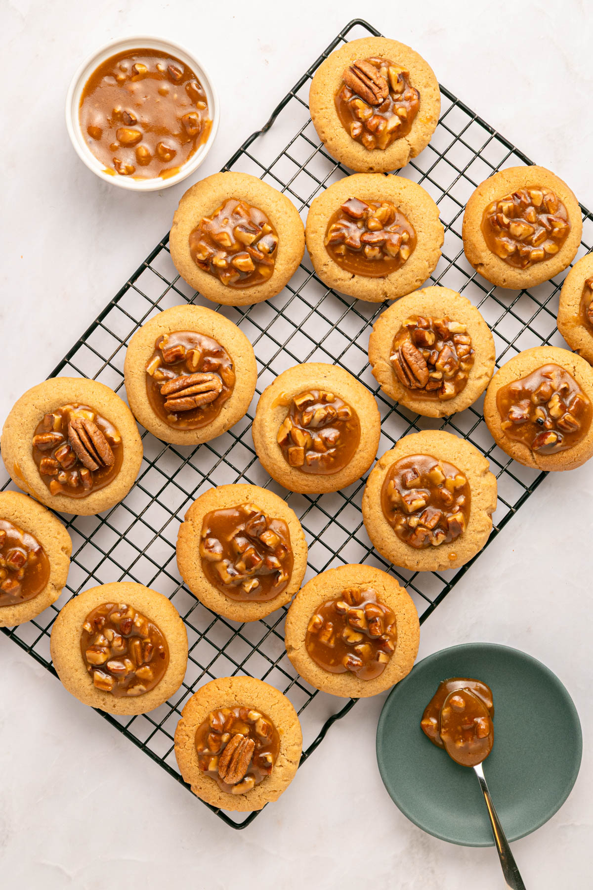A cooling rack holds several thumbprint cookies topped with caramel pecan filling. A bowl of extra filling and a spoon rest nearby on the table.