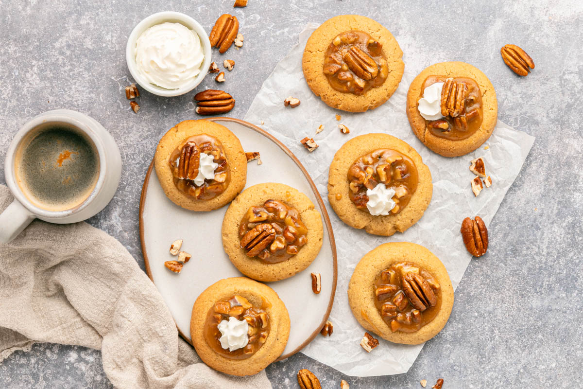 A plate of pecan cookies with caramel and whipped cream, surrounded by extra pecans, a cup of coffee, and a bowl of whipped cream on a grey surface.