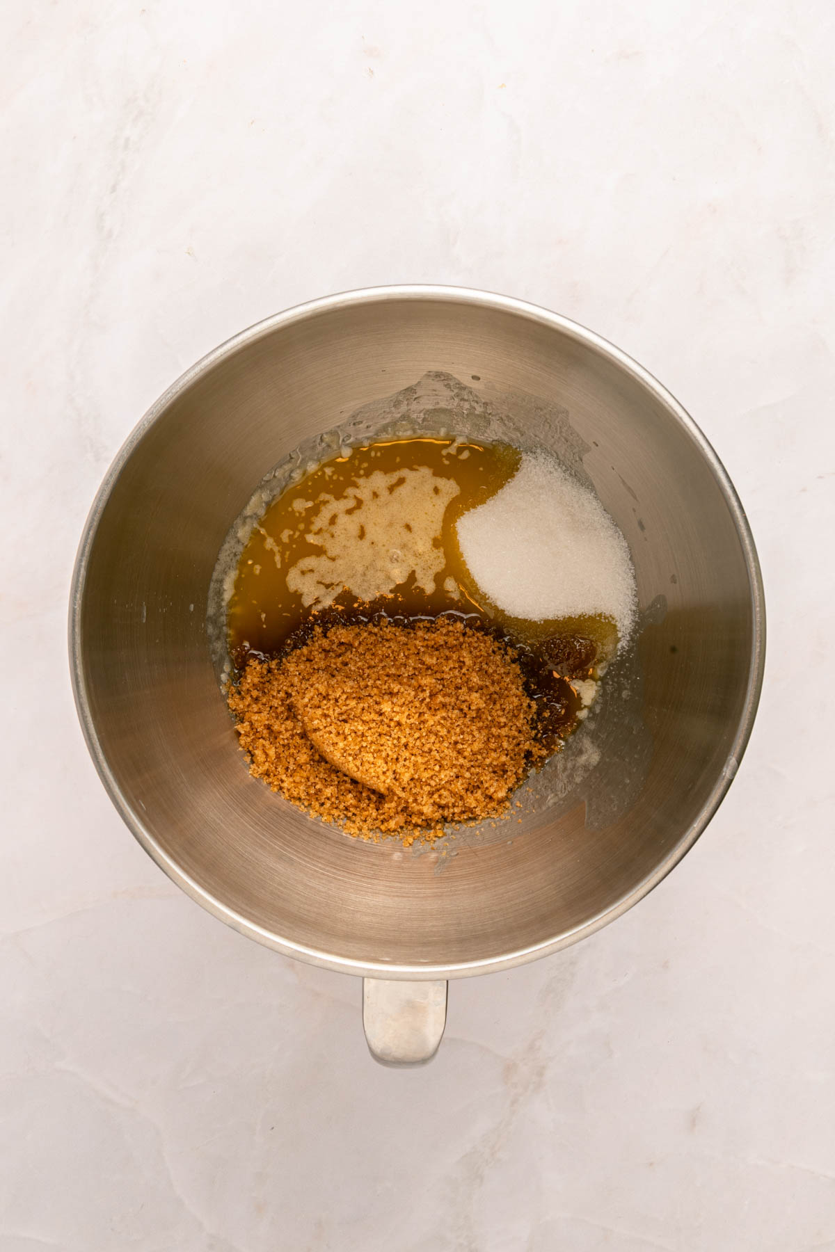 A metal mixing bowl containing brown sugar, granulated sugar, and melted butter on a light-colored surface.