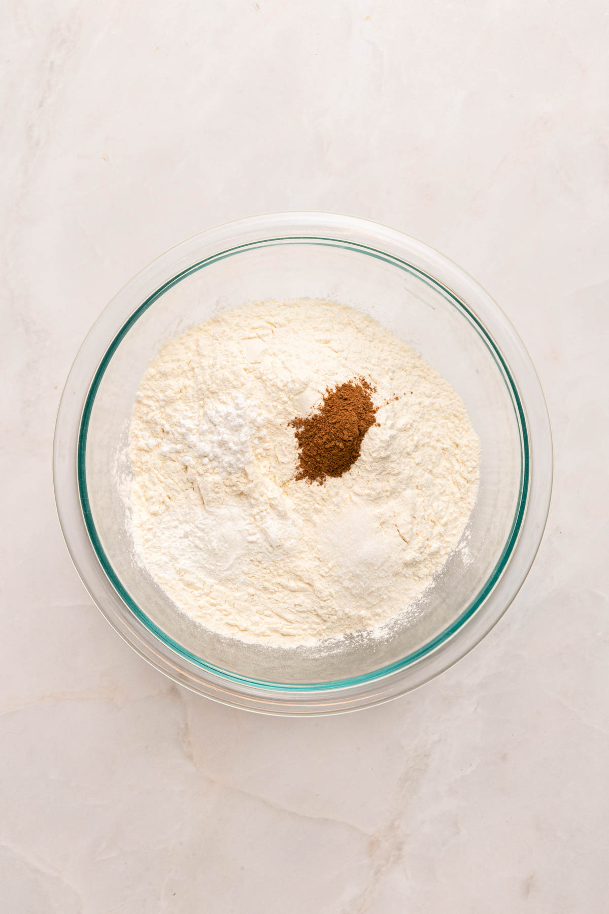 A glass bowl containing flour, baking powder, and a small amount of ground cinnamon on a light-colored surface.