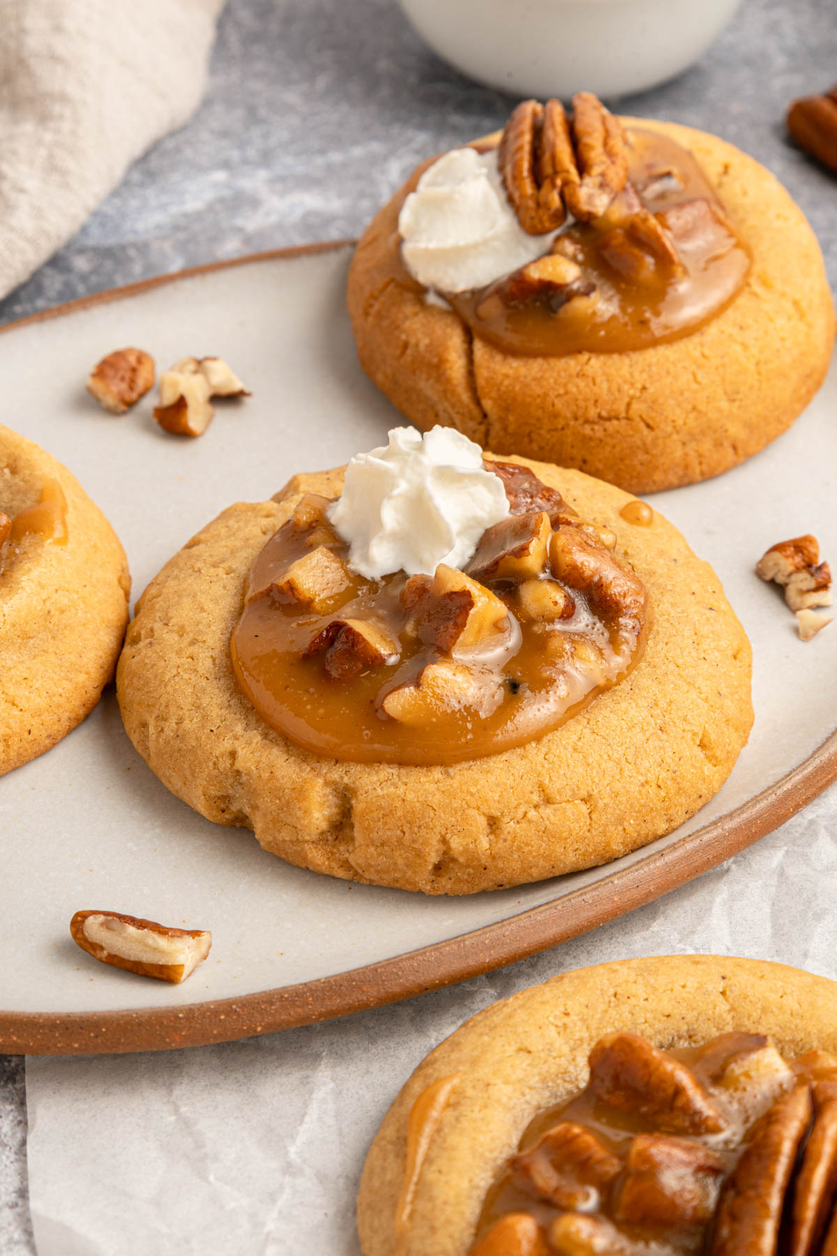 Three caramel pecan cookies topped with whipped cream and pecan pieces are arranged on a ceramic plate, with extra pecans scattered around.