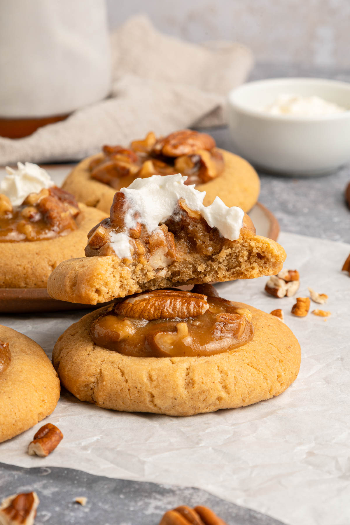 Two cookies topped with pecan pieces and cream, with one cookie broken in half to show the inside, placed on parchment paper with additional cookies and a bowl of cream in the background.