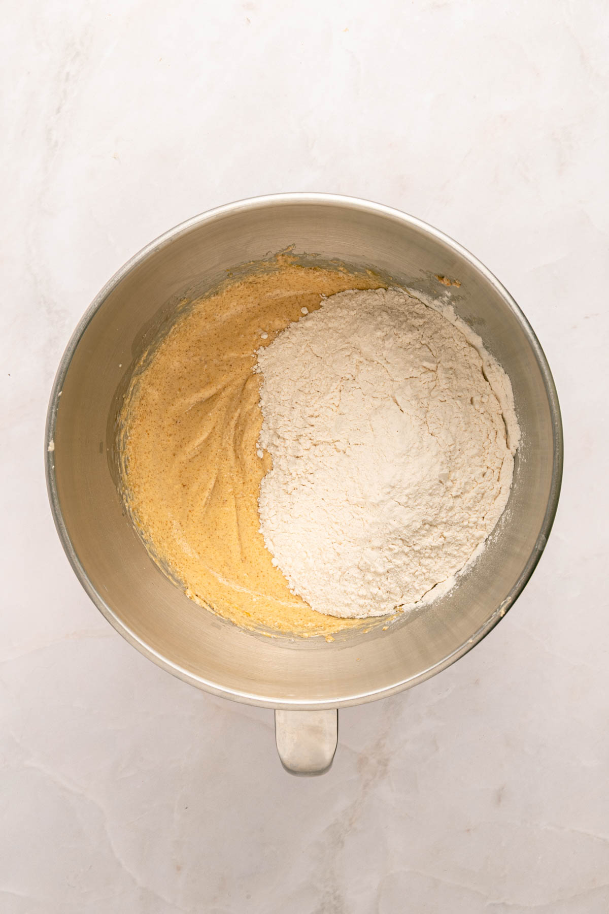 A mixing bowl containing partially mixed batter, with flour sitting on top of a yellowish mixture on one side.