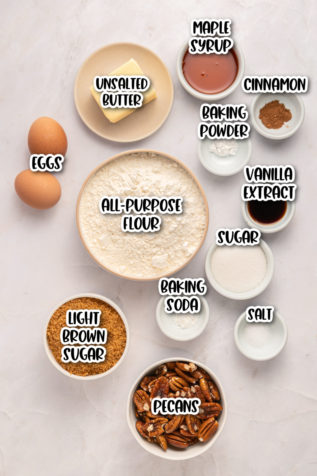A flat lay of labeled baking ingredients including eggs, unsalted butter, all-purpose flour, sugars, pecans, maple syrup, baking powder, baking soda, salt, cinnamon, and vanilla extract.