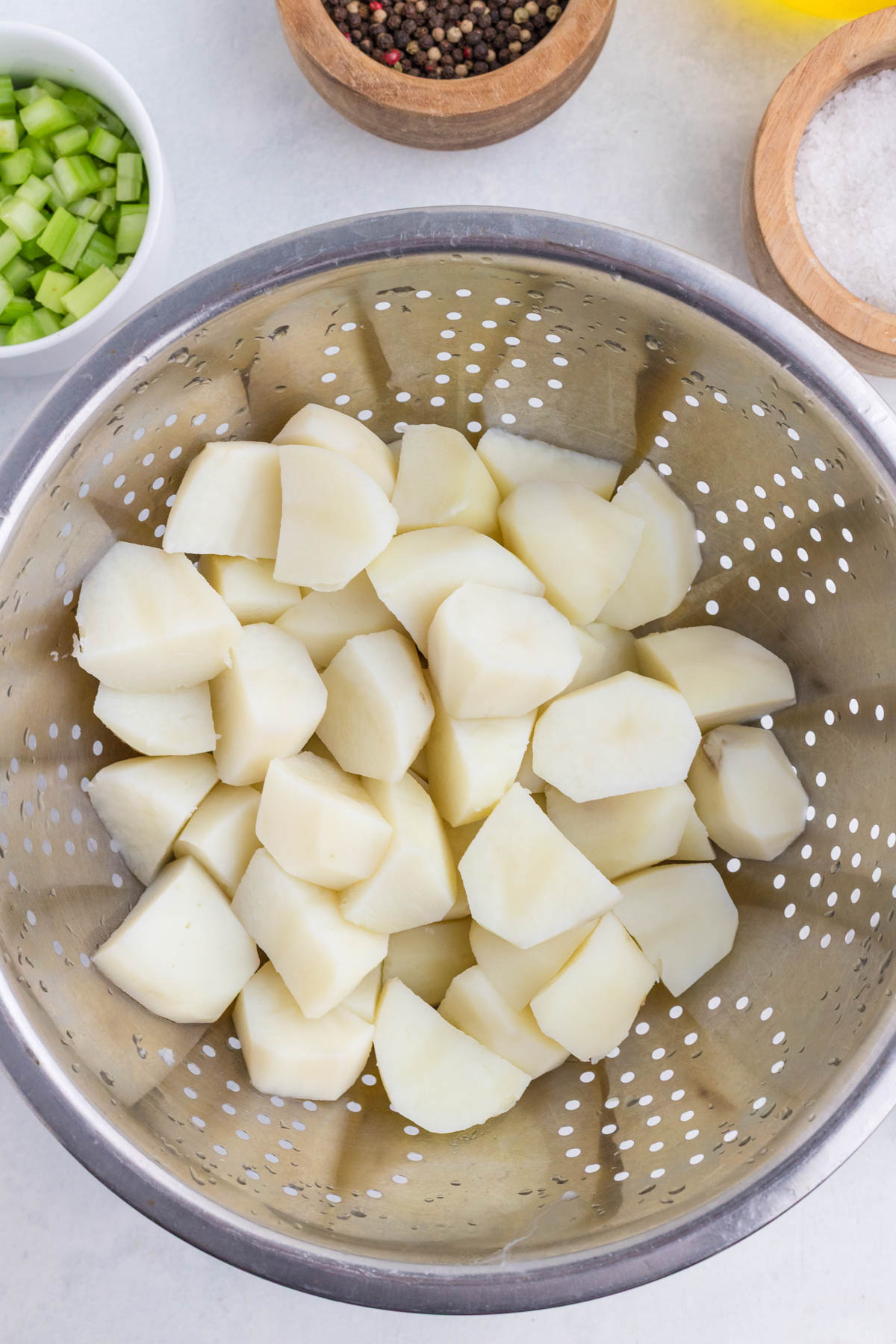 Peeled and chopped potatoes in a metal colander, with bowls of chopped celery, black peppercorns, and salt nearby.